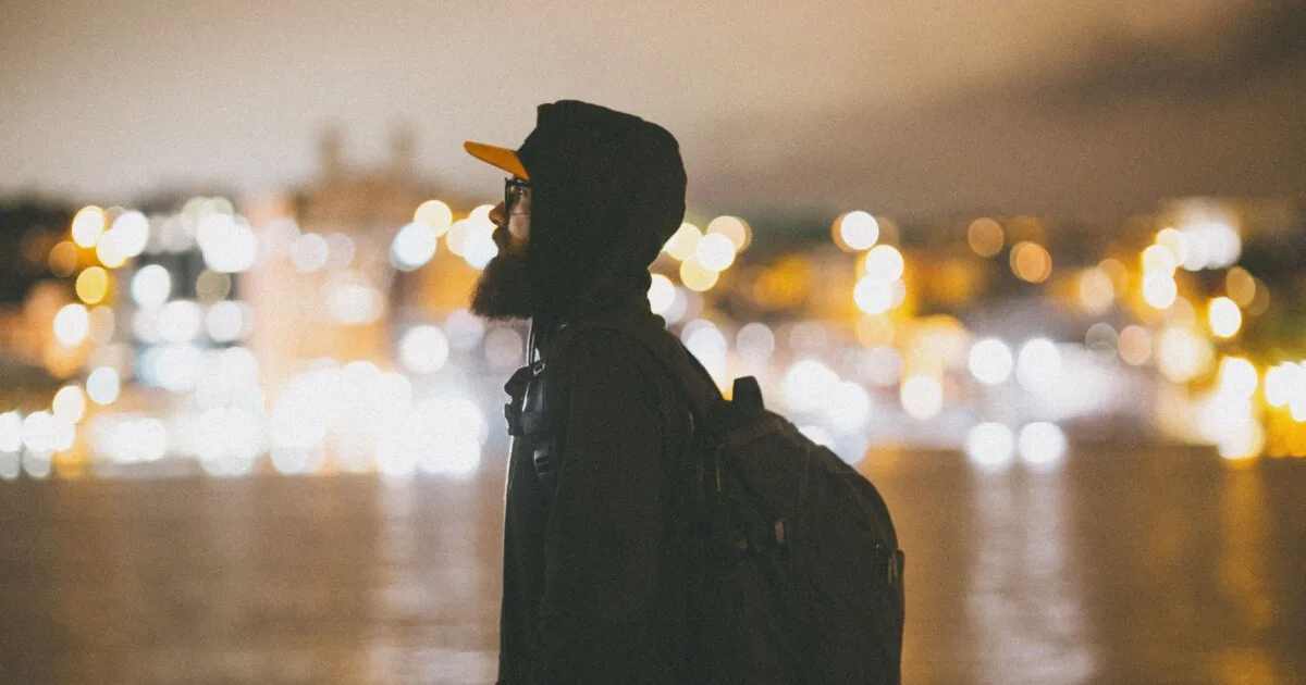 Man with backpack standing by city lights at night, gay solo travel in Lisbon