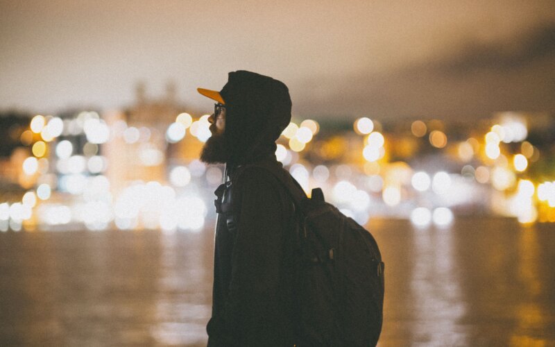 Man with backpack standing by city lights at night, gay solo travel in Lisbon